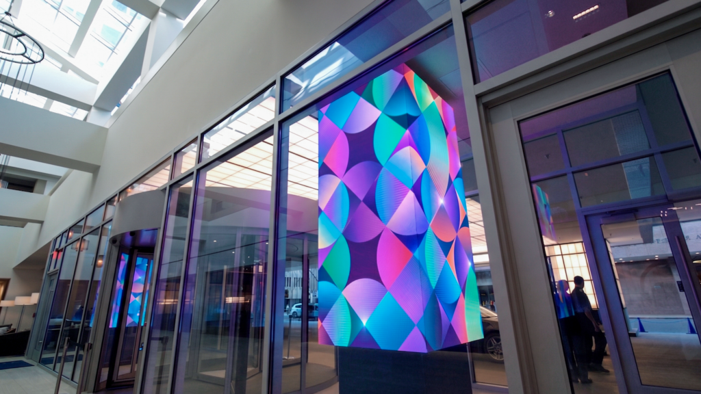 A multi-sided LED pillar displaying vibrant, colorful geometric patterns inside a modern glass-walled atrium venue.