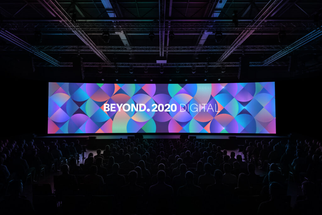 A wide view of a darkened conference hall filled with an audience seated in silhouettes. Stage-wide LED screen displays colorful, overlapping geometric circle patterns with the text "BEYOND.2020 DIGITAL" centered in white.